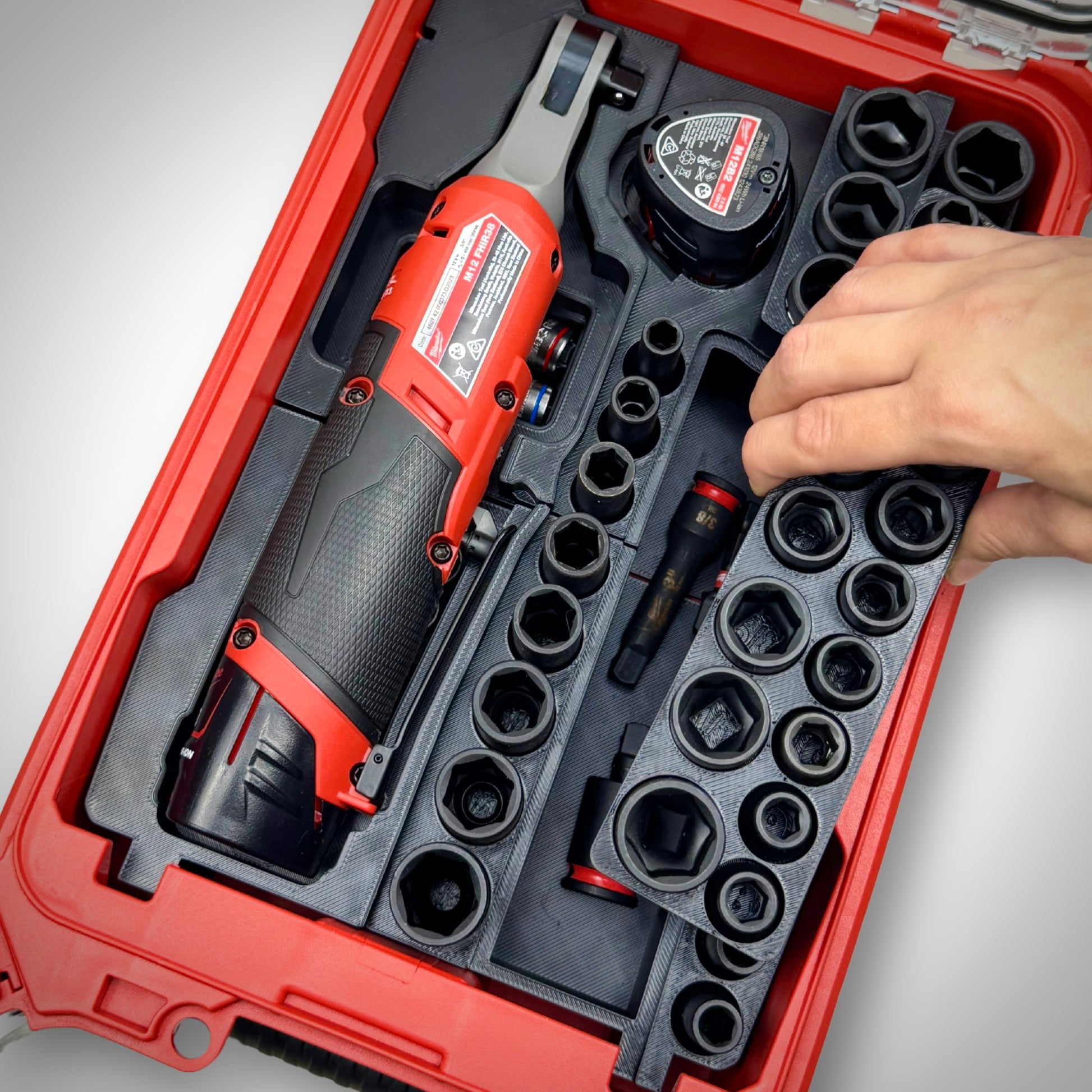 Hand lifting a removable storage tray from the M12 Ratchet & 3/8" Socket Set Insert inside the Milwaukee PACKOUT™ Compact Organiser, revealing hidden storage compartments underneath.